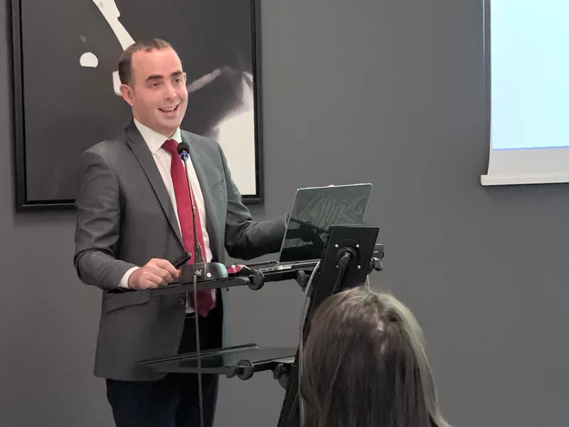 Koyu renk takım elbise ve kırmızı kravatla, dizüstü bilgisayarlı kürsüde sunum yapan Prof. Dr. Yusuf Yıldırım; arkada gri duvar.