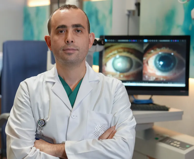 Muayenehanede beyaz önlükle, kollarını kavuşturmuş Prof. Dr. Yusuf Yıldırım; arkadaki ekranda iki göz görüntüsü.