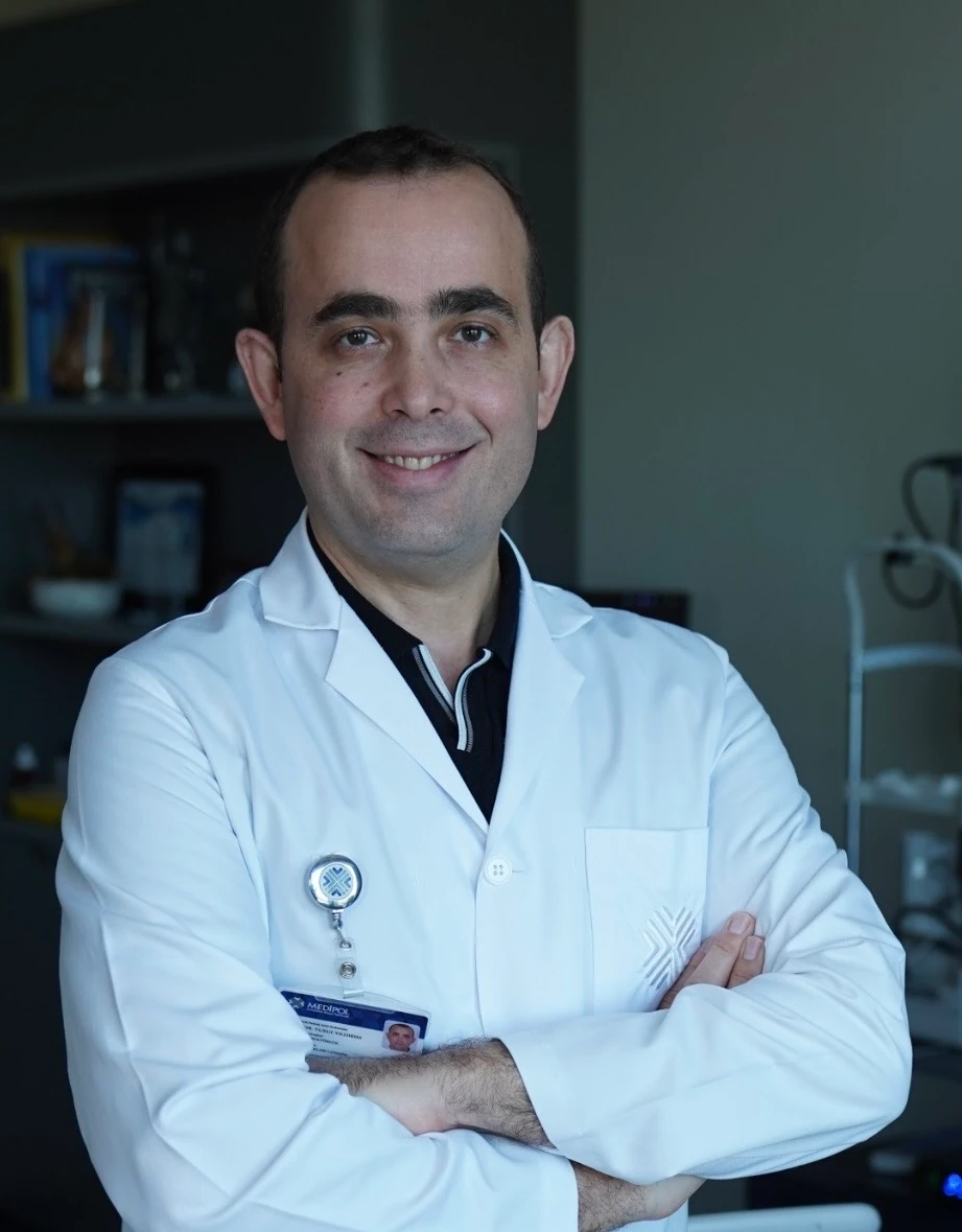 Prof. Dr. Yusuf Yıldırım beyaz önlük ve boynunda kimlik kartıyla, kollarını kavuşturmuş, gülümsüyor.